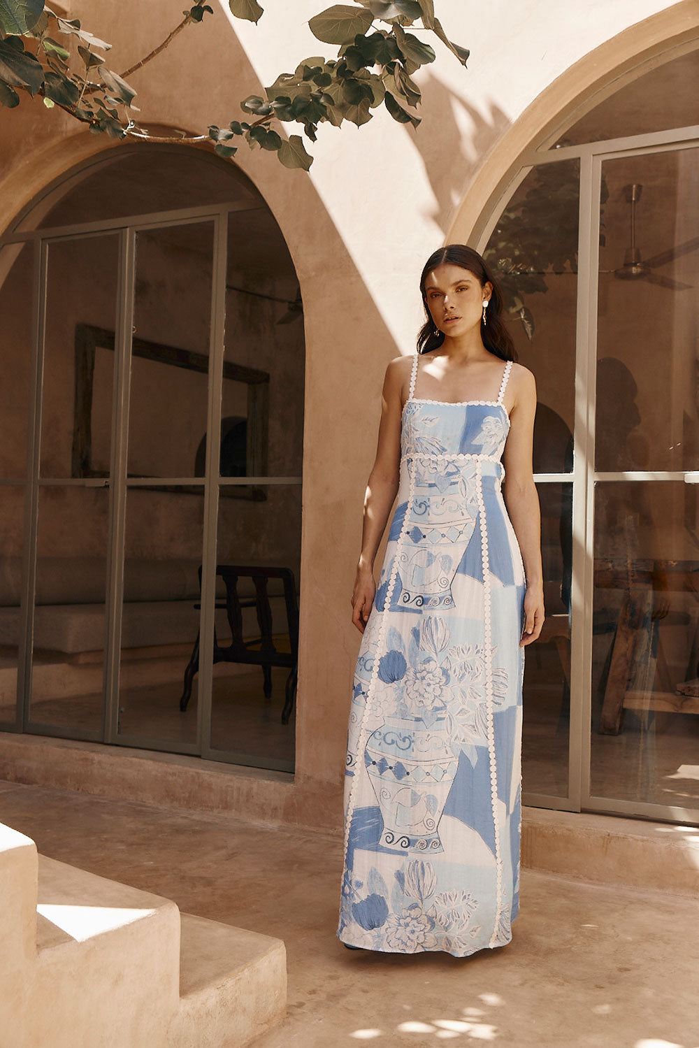 Model standing in a sunlit courtyard wearing a blue and white vase print maxi dress with white ric rac trim