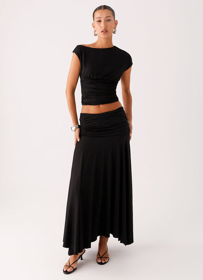 Model wearing a black low-rise ruched maxi skirt with a flared hem and matching cap sleeve top