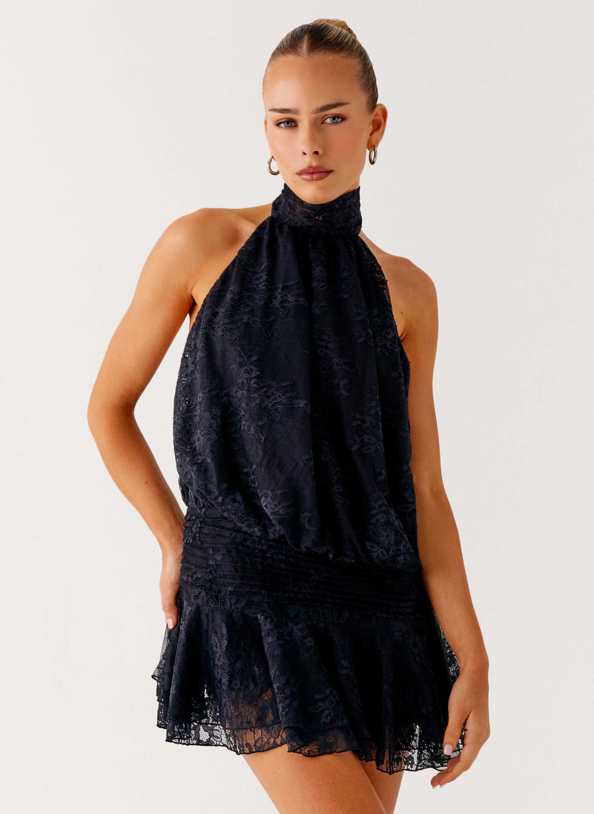 Model wearing a black lace halter neck mini dress with a pleated drop waist and tiered flared hem