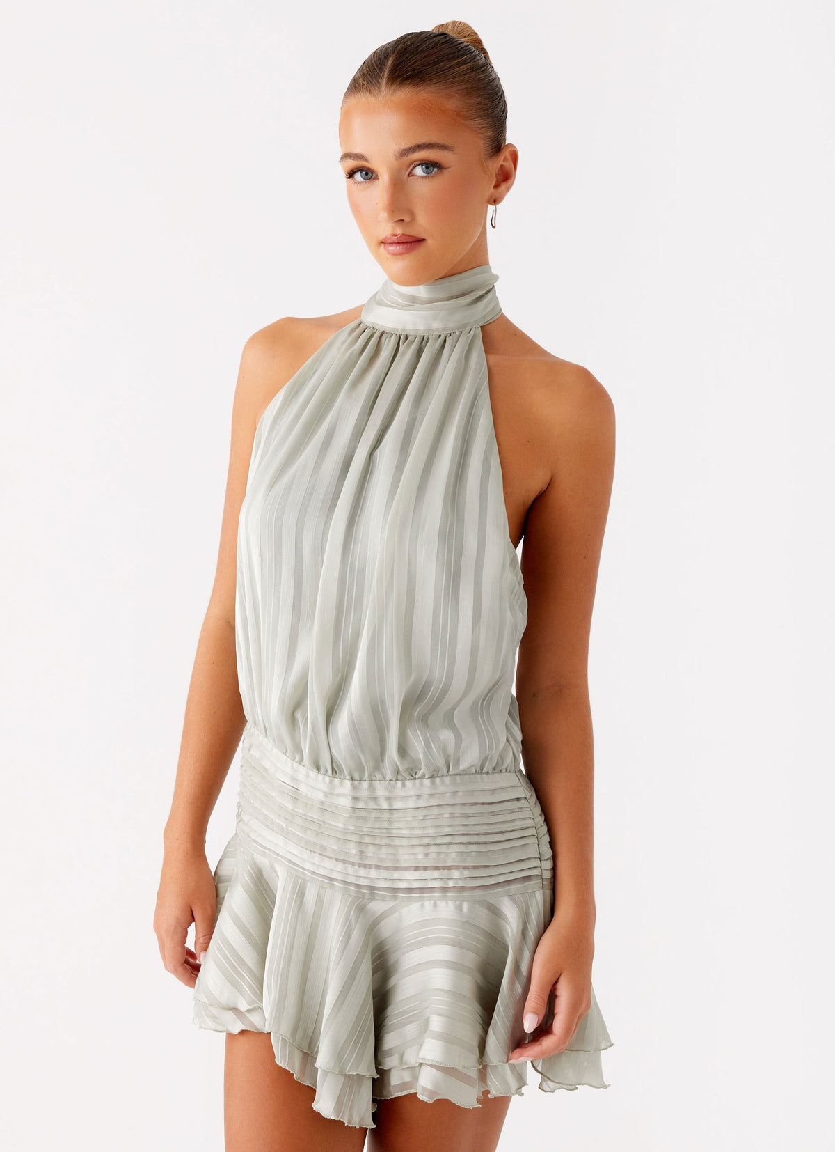 Model wearing a sage green chiffon halter neck mini dress with a pleated drop waist and flared hem