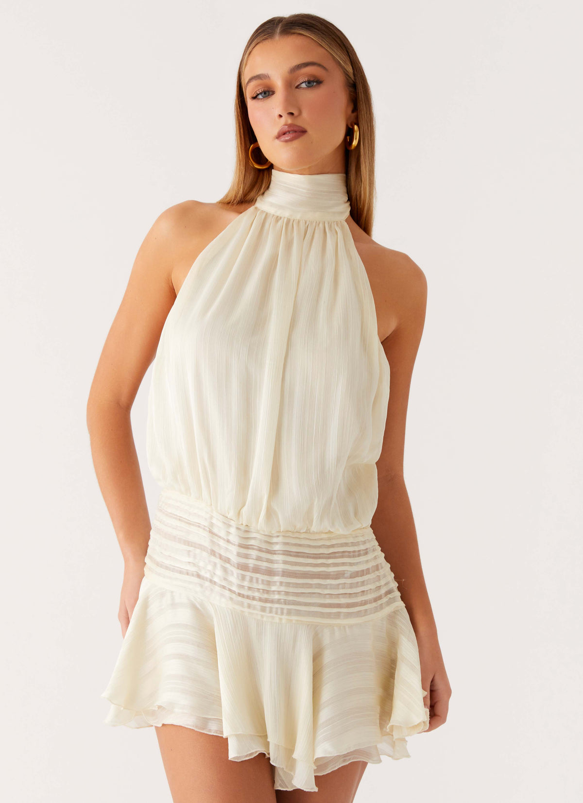 Model wearing an ivory chiffon halter neck mini dress with a pleated drop waist and tiered flared hem
