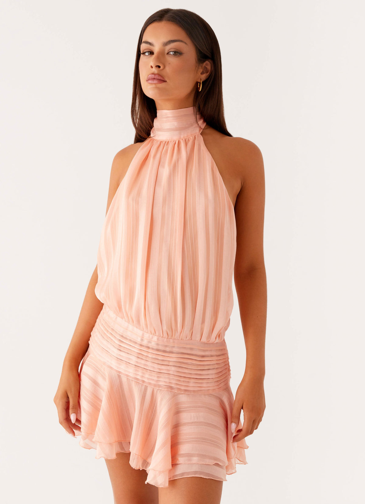 Model wearing a peach chiffon halter neck mini dress with a pleated drop waist and tiered flared hem