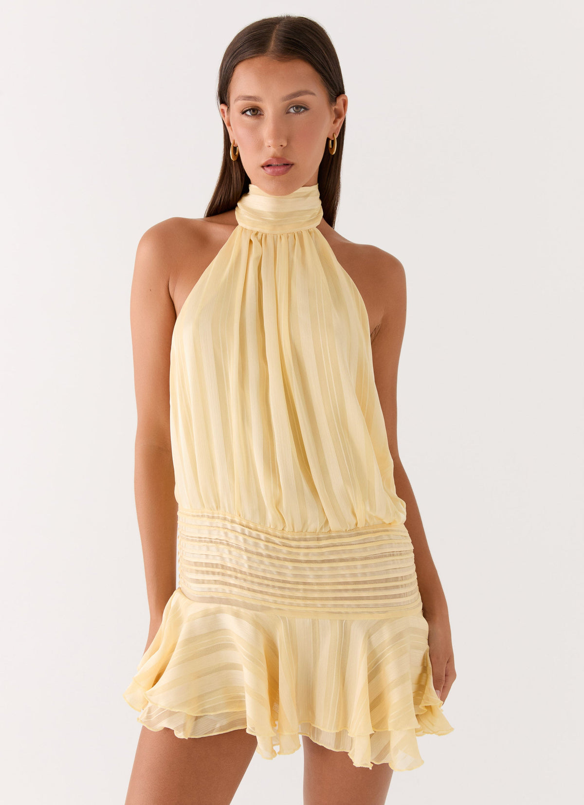 Model wearing a pale yellow chiffon halter neck mini dress with a pleated drop waist and flared hem